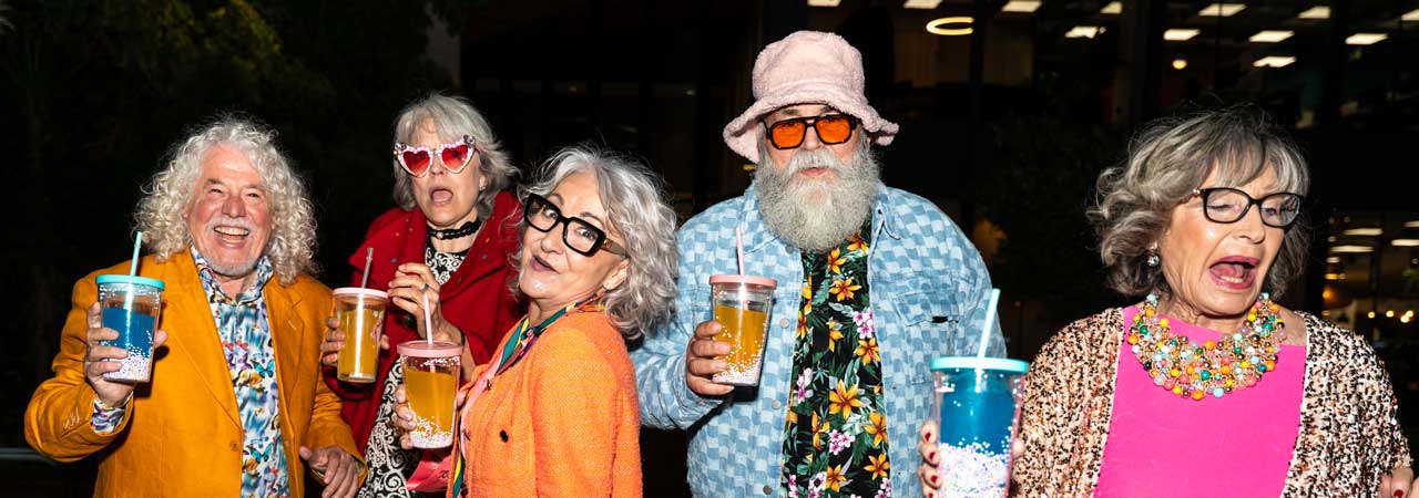 Gruppe lebensfroher Senioren auf einer Party, fröhlich lachend in farbenfrohen Outfits mit Getränken in der Hand. Symbol für aktives, modernes und glückliches Leben im Alter.